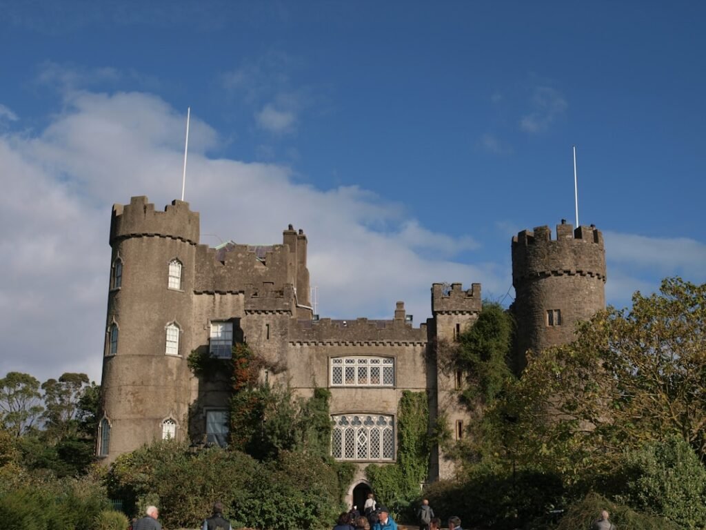 a group of people standing in front of a Fachada do Malahide Castle com gramado verde e árvores floridas ao redor, céu parcialmente azul